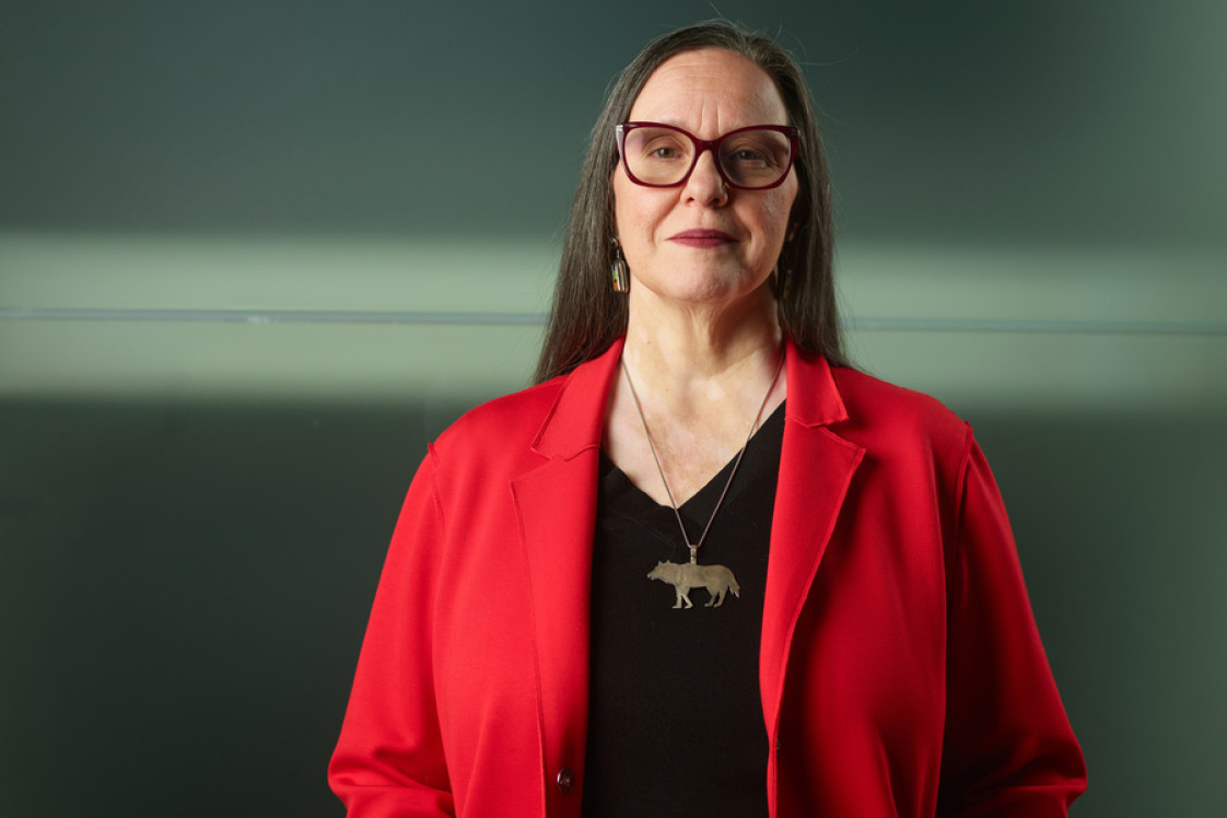 Professor Sonya Atalay stand in red jacket and wolf necklace against a grey and green background