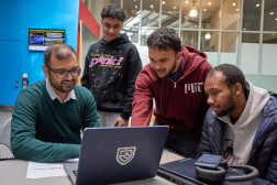 Four MIT students look at a laptop together in a large open space